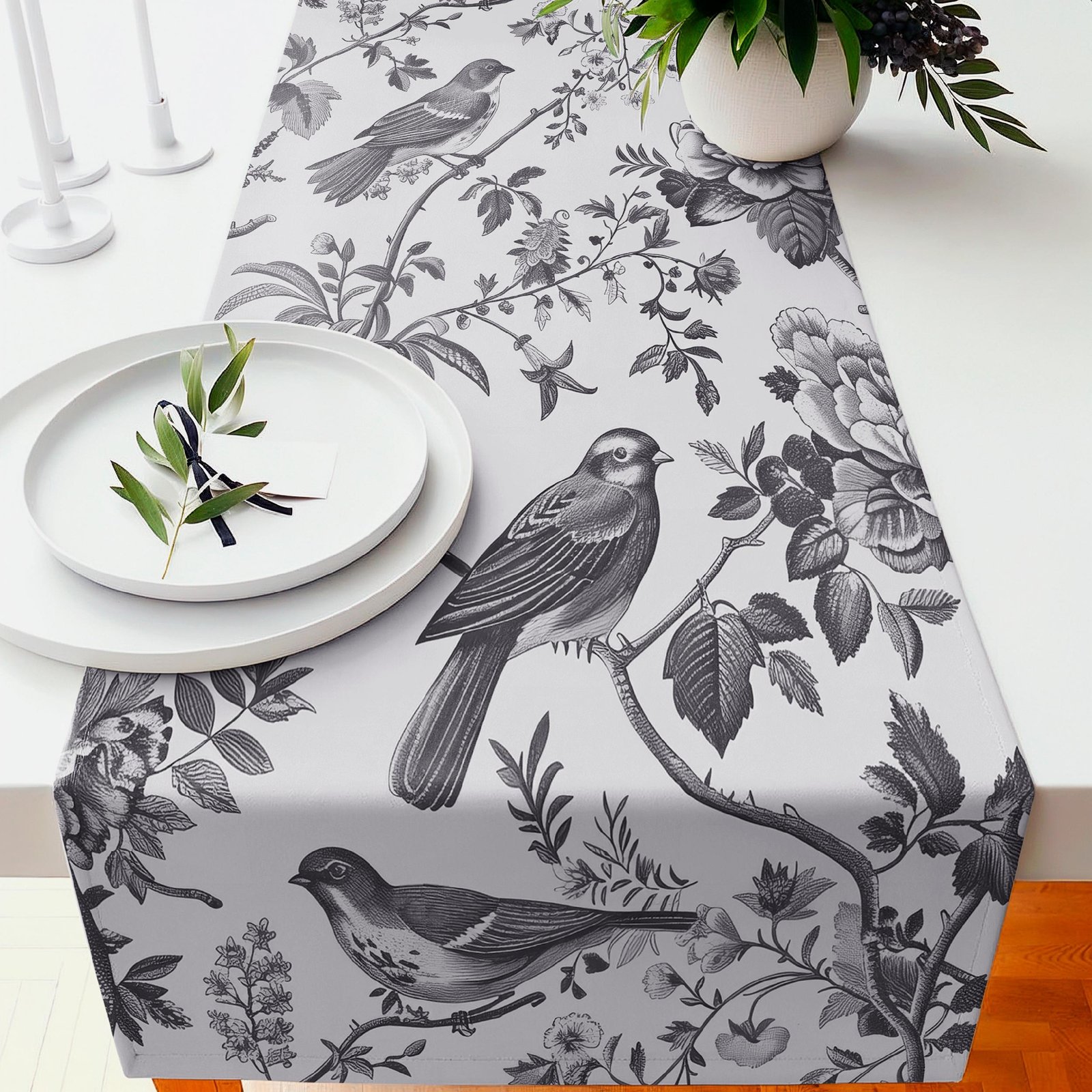 Close-up of a white sofa or chair covered in a black botanical print with birds, next to a white vase of greenery and candles on the left.